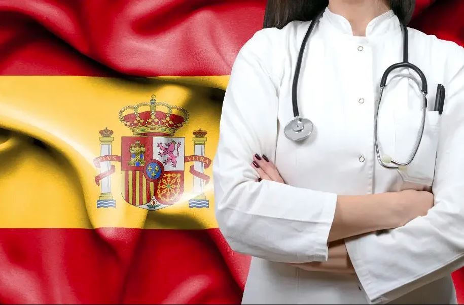 Doctor in a white coat with a stethoscope, standing with arms crossed, against the backdrop of the Spanish flag.