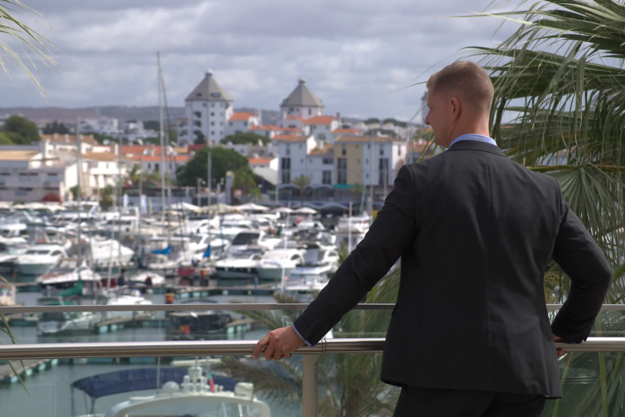 Man in a suit standing on a balcony overlooking a marina filled with boats, with buildings and cloudy skies in the background.