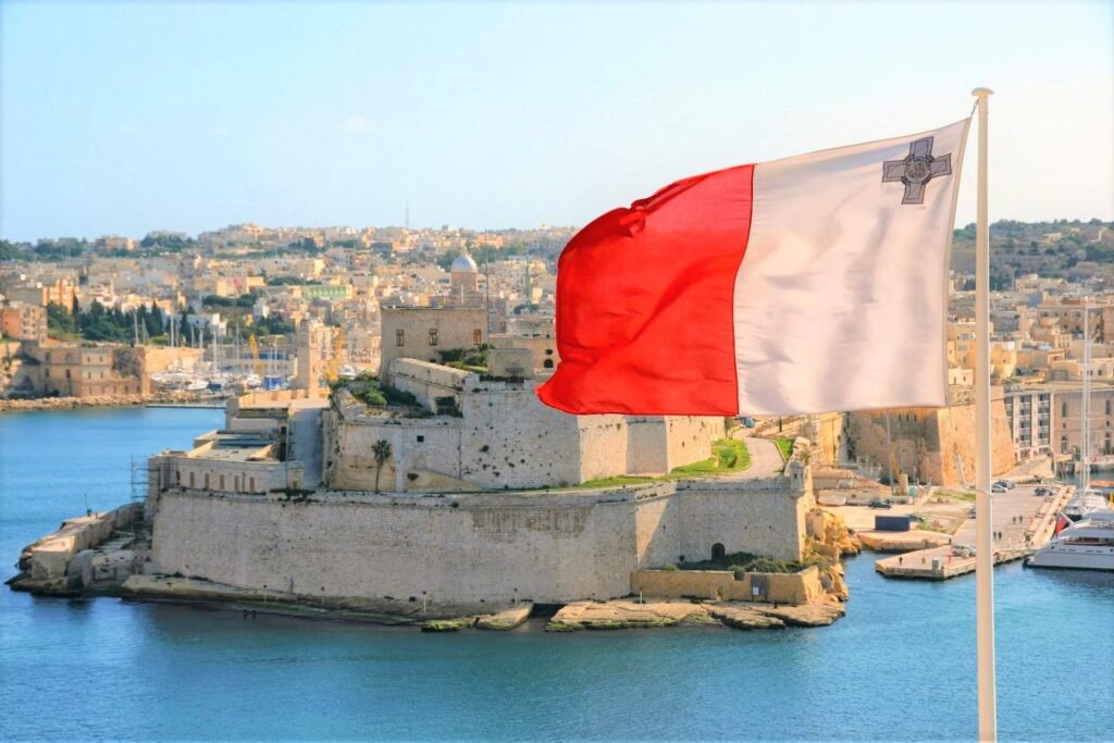 Maltese flag waving in the foreground with a historic stone fortress and a harbor surrounded by buildings in the background under a clear sky.