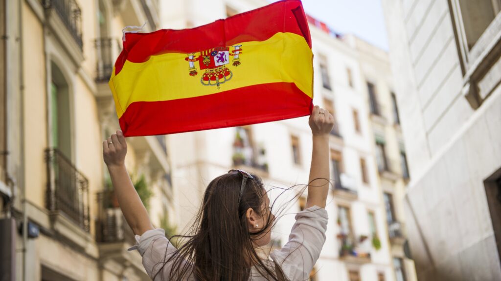 Woman holding up the Spanish flag in an urban setting with buildings and balconies in the background.