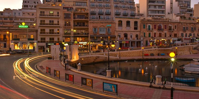 City street at dusk with light trails from passing cars, a small waterfront area, and buildings illuminated by warm lights in the background.